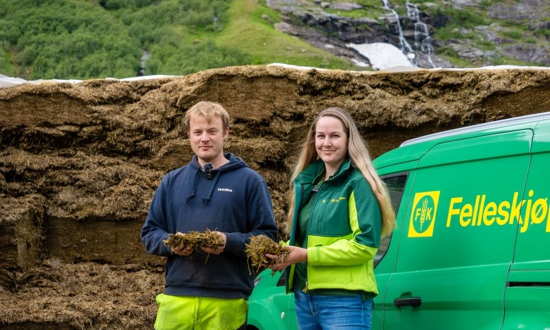 To personer i en plansilo holder grovfôr med bil i bakgrunnen.