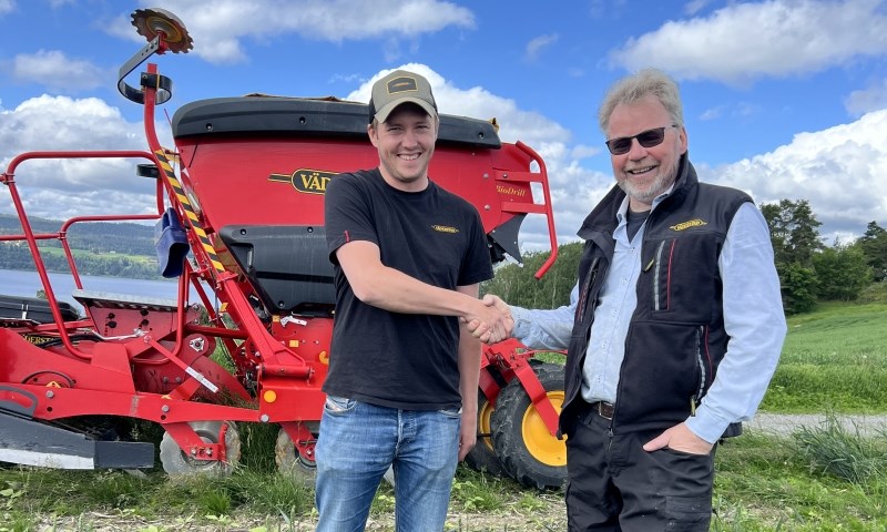 Kornåker med såmaskin og to karer med himmel og vann i bakgrunnen