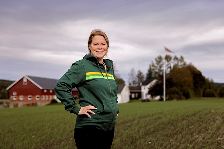 Smilende kvinne på et jorde, med et gårdstun, med rød driftsbygning og hvitt hovedhus i bakgrunnen.